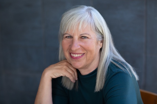 Close up picture of Judy Kahn siting in a chair, smiling and wearing a black blouse with her hand nested under her chin while she is looking at the camera.